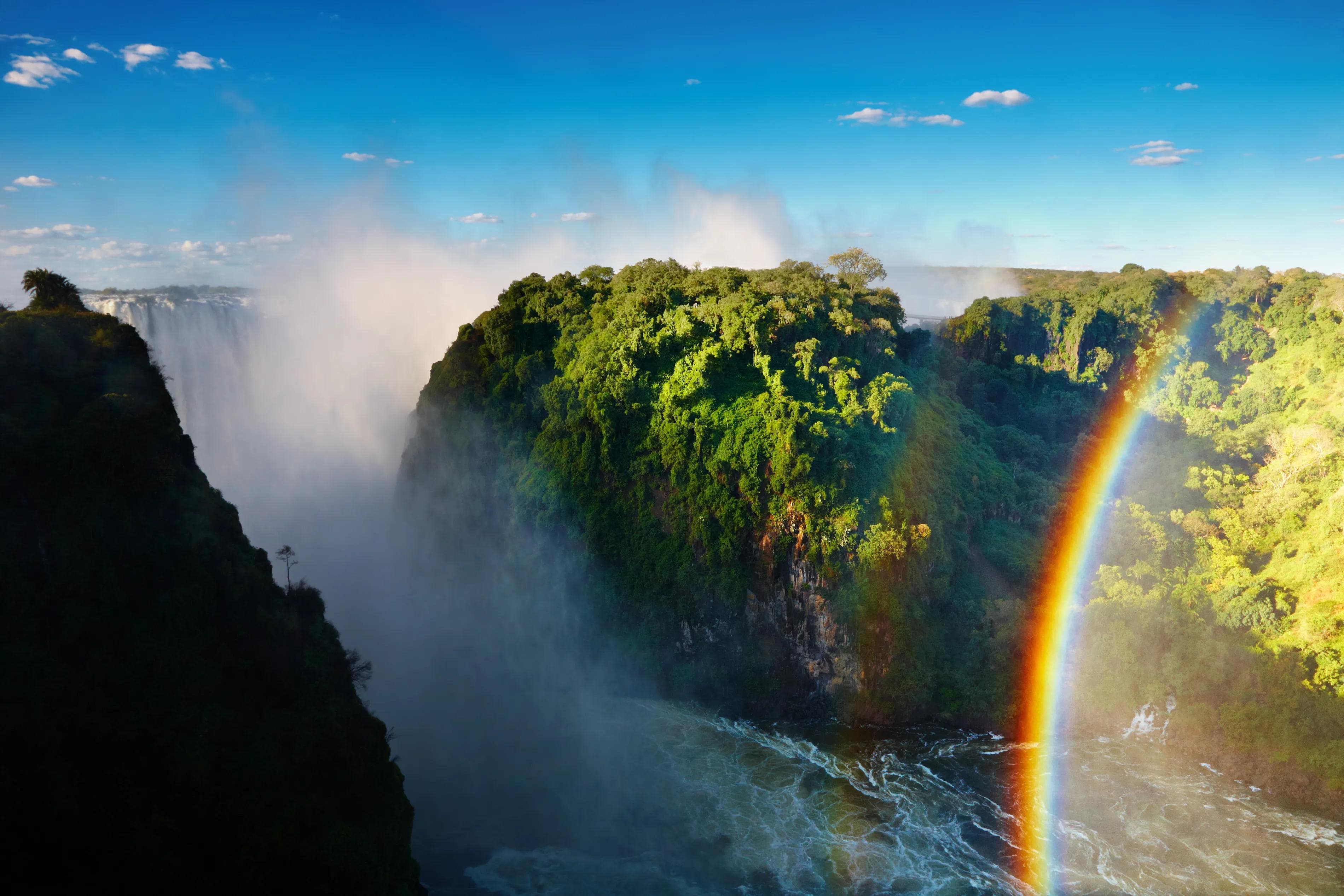 Victoria Falls with mist rising over the gorge and a rainbow arching through the spray, viewed from the Zimbabwe side